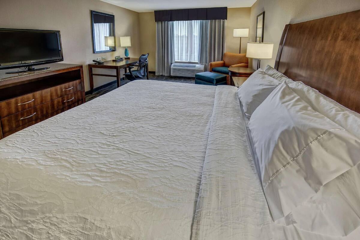 A hotel room with a large bed, a desk with a computer, a television, a chair, and a window with curtains.