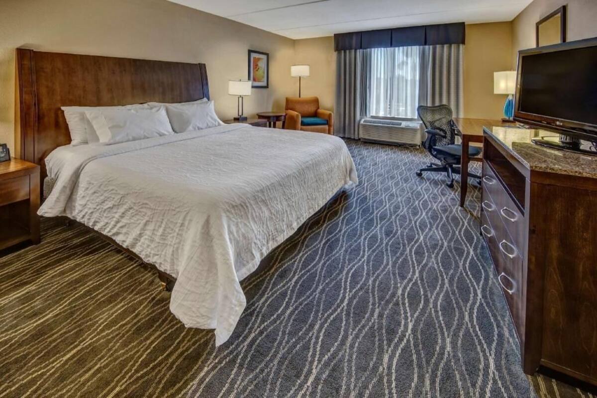 A hotel room with a large bed, a desk with a flat-screen TV, a chair, a desk lamp, and a window with curtains.