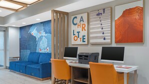Un espace de bureau moderne avec un canapé bleu, des chaises orange, un bureau doté de deux écrans, une imprimante et des œuvres d’art murales.