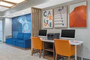 A modern office space with a blue sofa, orange chairs, a desk with two monitors, a printer, and wall art. - Holiday Inn Express Hotel & Suites Charlotte by IHG (Charlotte)
