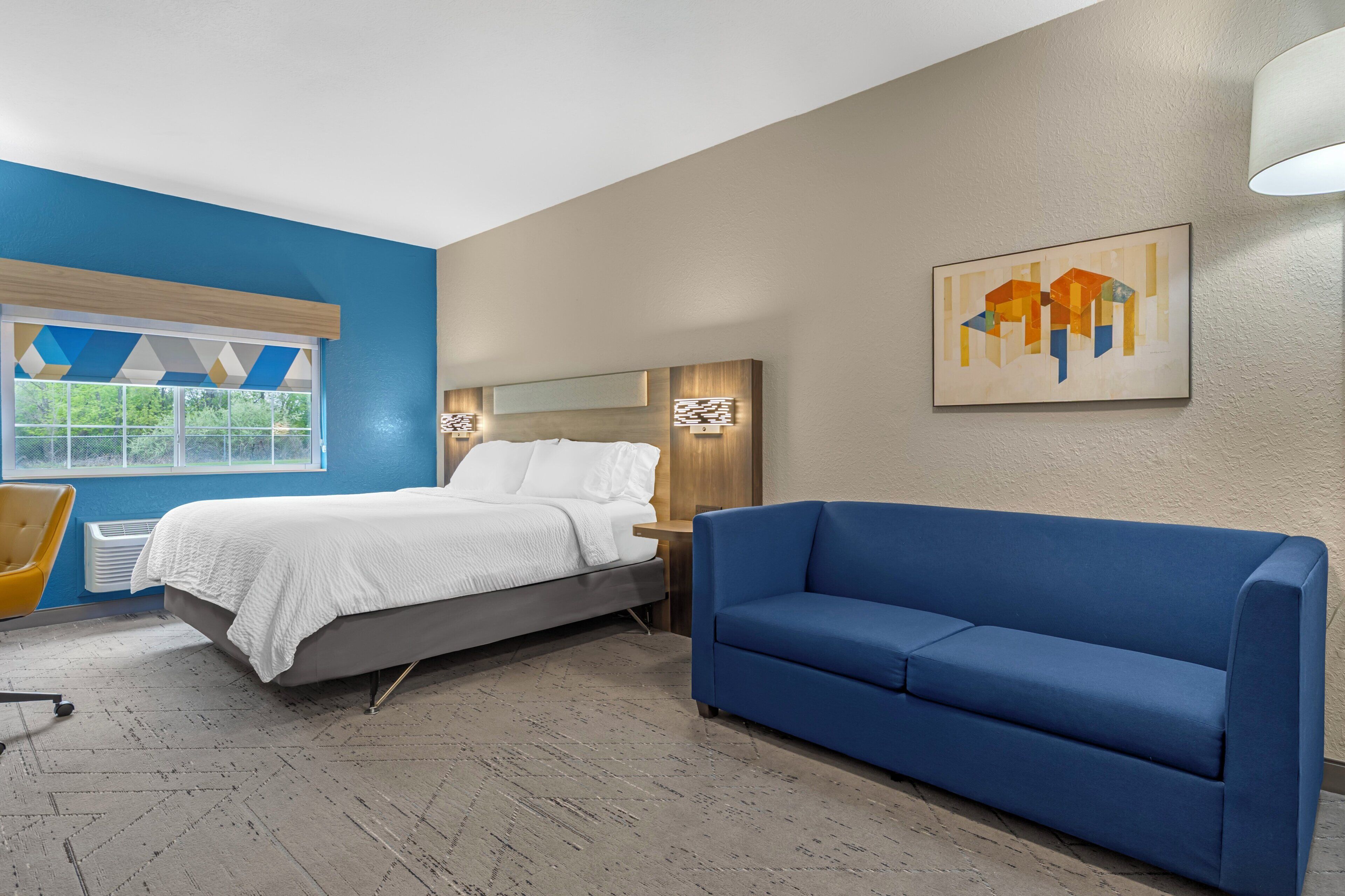 A hotel room with a bed, a blue sofa, a chair, and a painting on the wall.