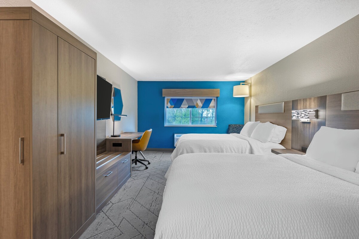 Une chambre d’hôtel comprenant un lit, un bureau, une chaise, une armoire et une fenêtre donnant sur des arbres.