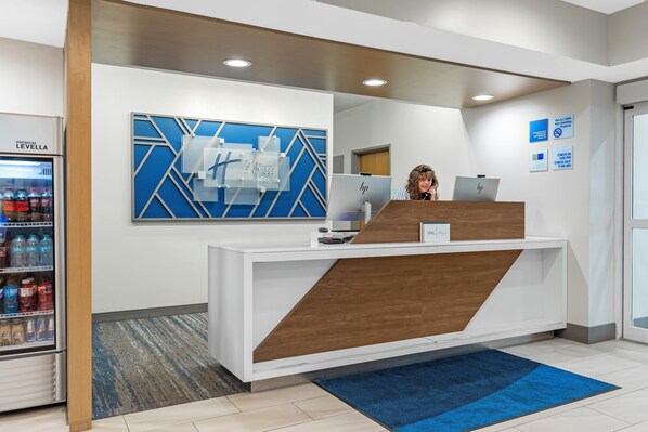 A reception desk with a blue geometric wall art, a refrigerator with beverages, and a blue carpet. - Holiday Inn Express Hotel & Suites Charlotte by IHG (Charlotte)