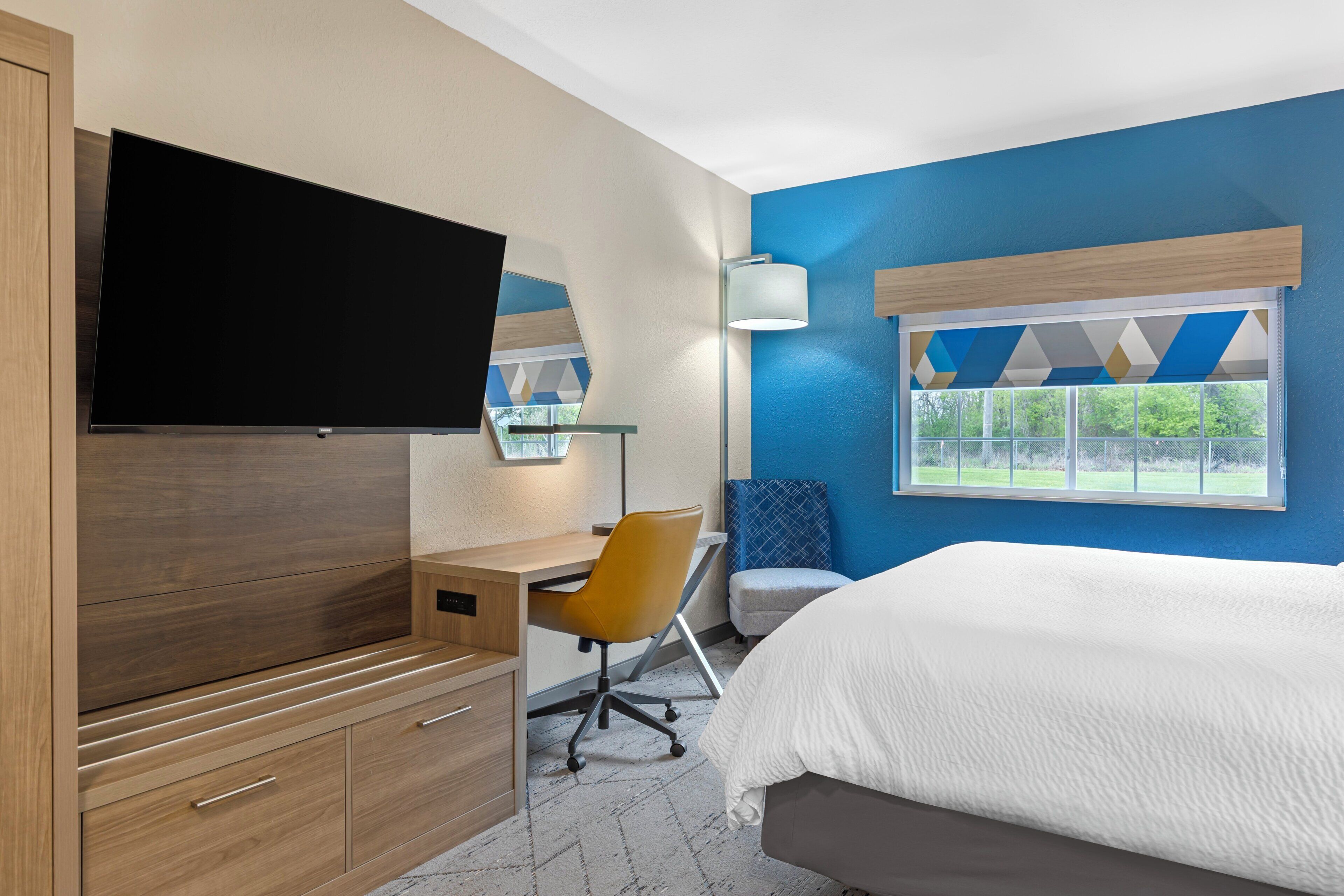 A hotel room with a bed, desk, chair, TV, window, and a lamp.