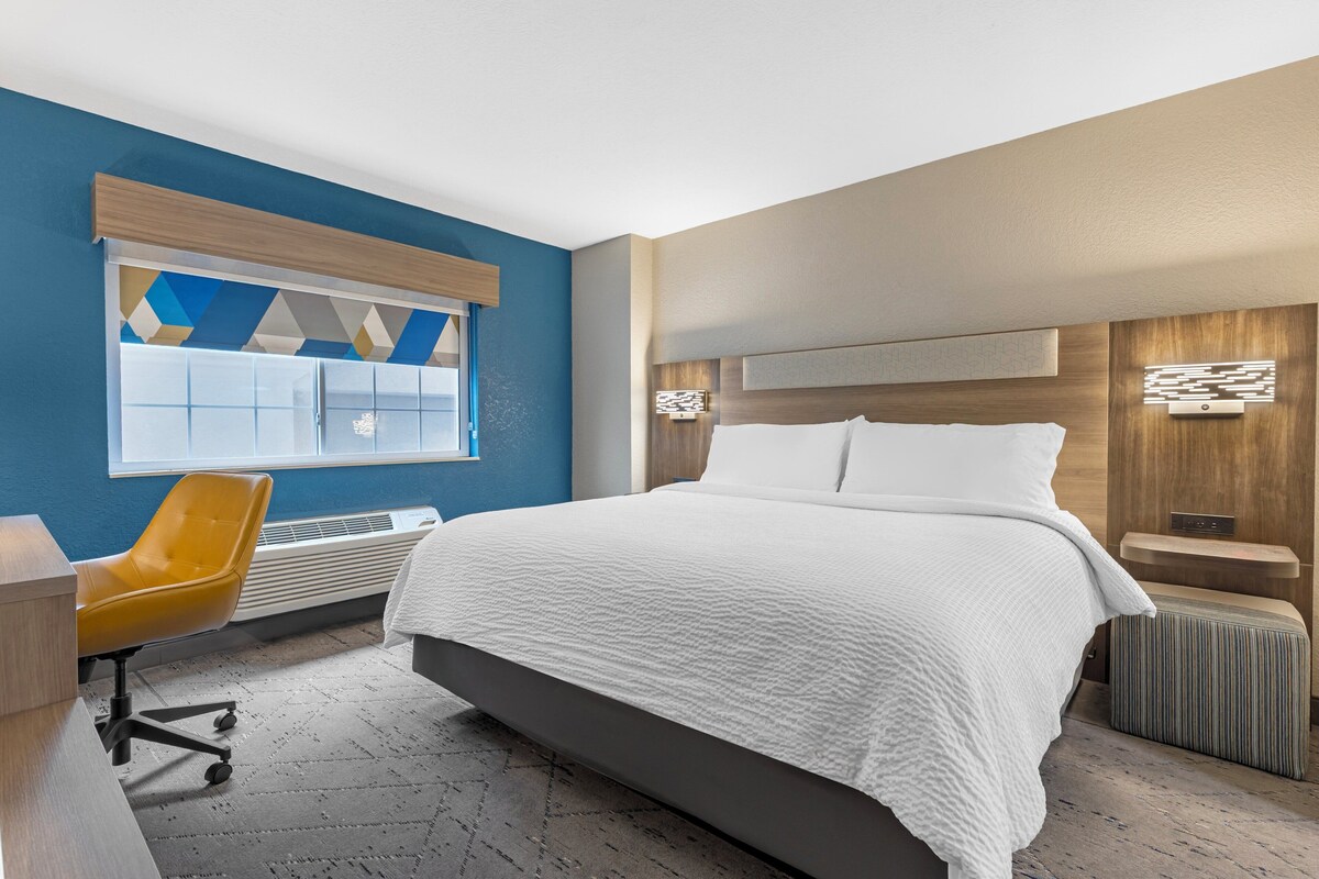 Une chambre d’hôtel moderne, dotée d’un grand lit, d’un bureau, d’une chaise et d’une fenêtre avec des stores à motifs géométriques.