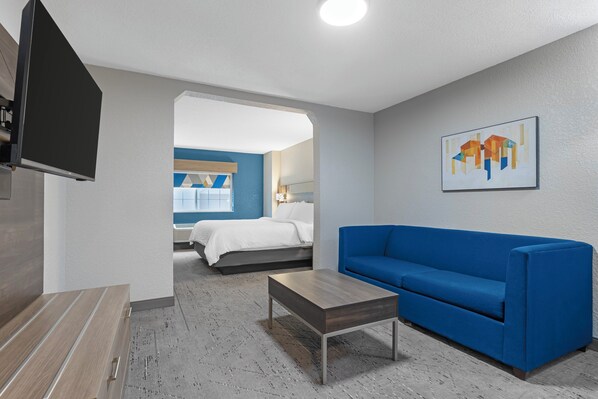 A modern hotel room with a blue sofa, a wooden coffee table, a flat-screen TV, and a painting on the wall. - Holiday Inn Express Hotel & Suites Charlotte by IHG (Charlotte)
