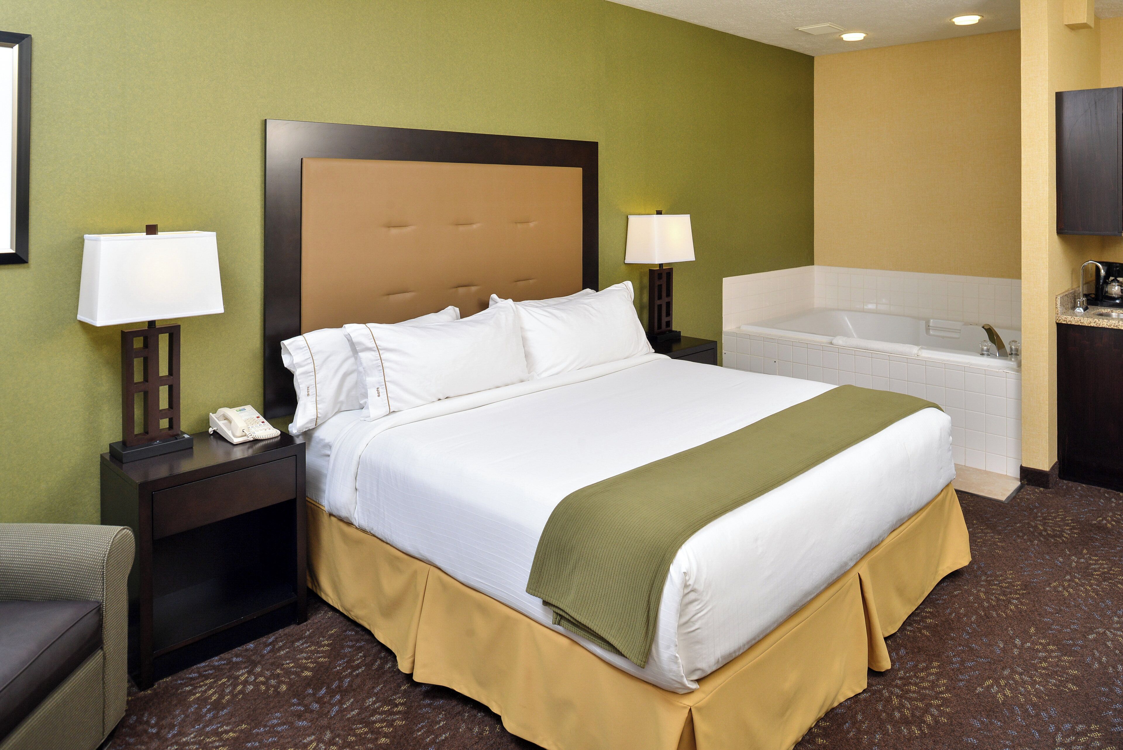 A hotel room with a large bed, a bedside table with a lamp, a telephone, and a bathroom with a bathtub and sink.