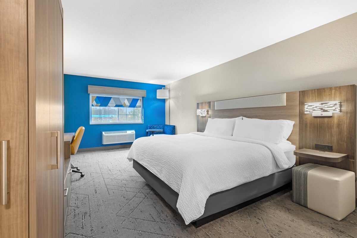 Une chambre d’hôtel moderne avec un grand lit, une table de chevet, une lampe et une fenêtre avec des stores.