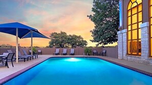 Piscina com espreguiçadeiras e um guarda-sol azul, cercada por um muro de pedra e árvores, com um pôr do sol ao fundo.
