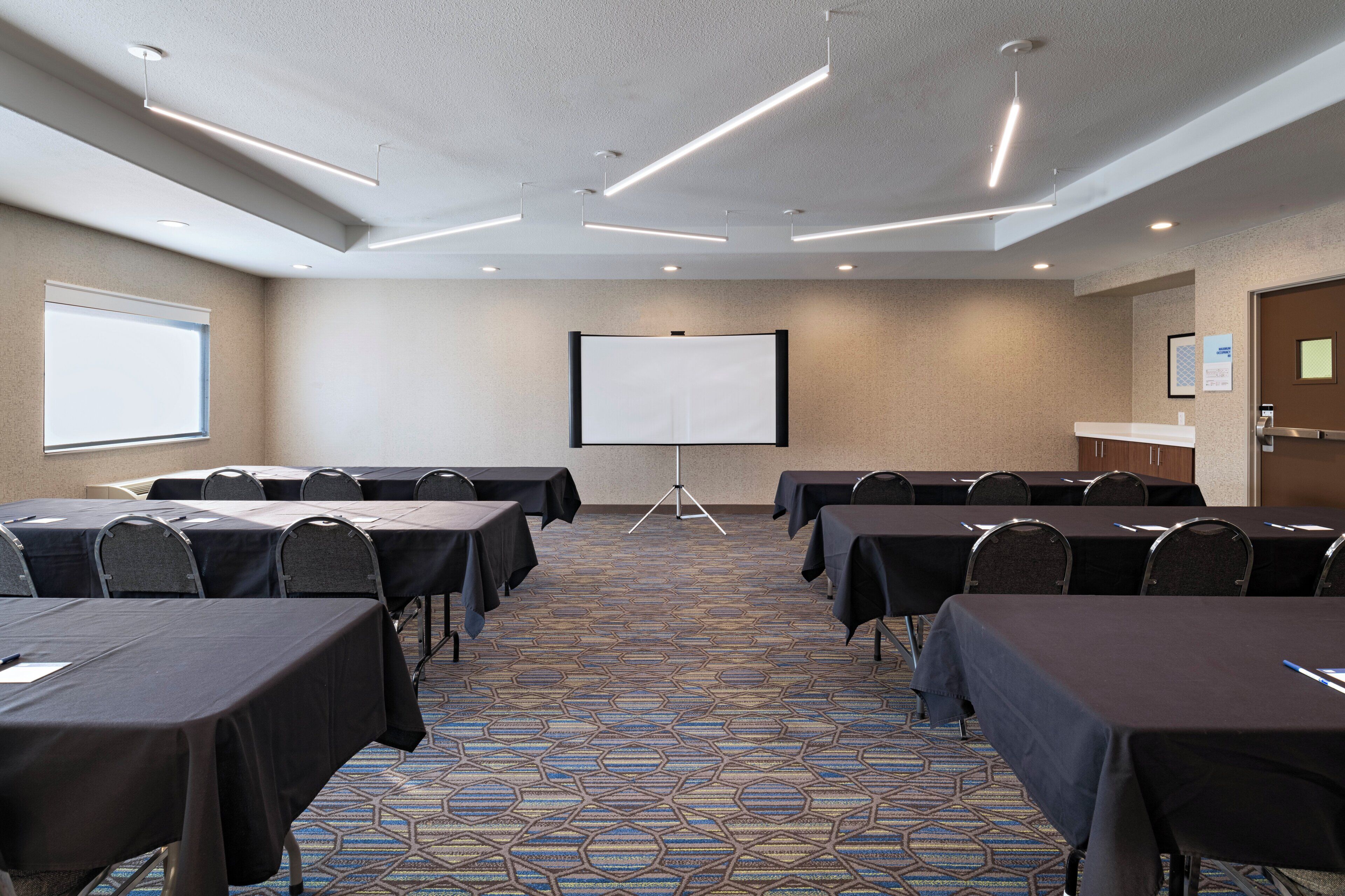 Un salón de conferencias con mesas, sillas, una pantalla de proyector y piso alfombrado.