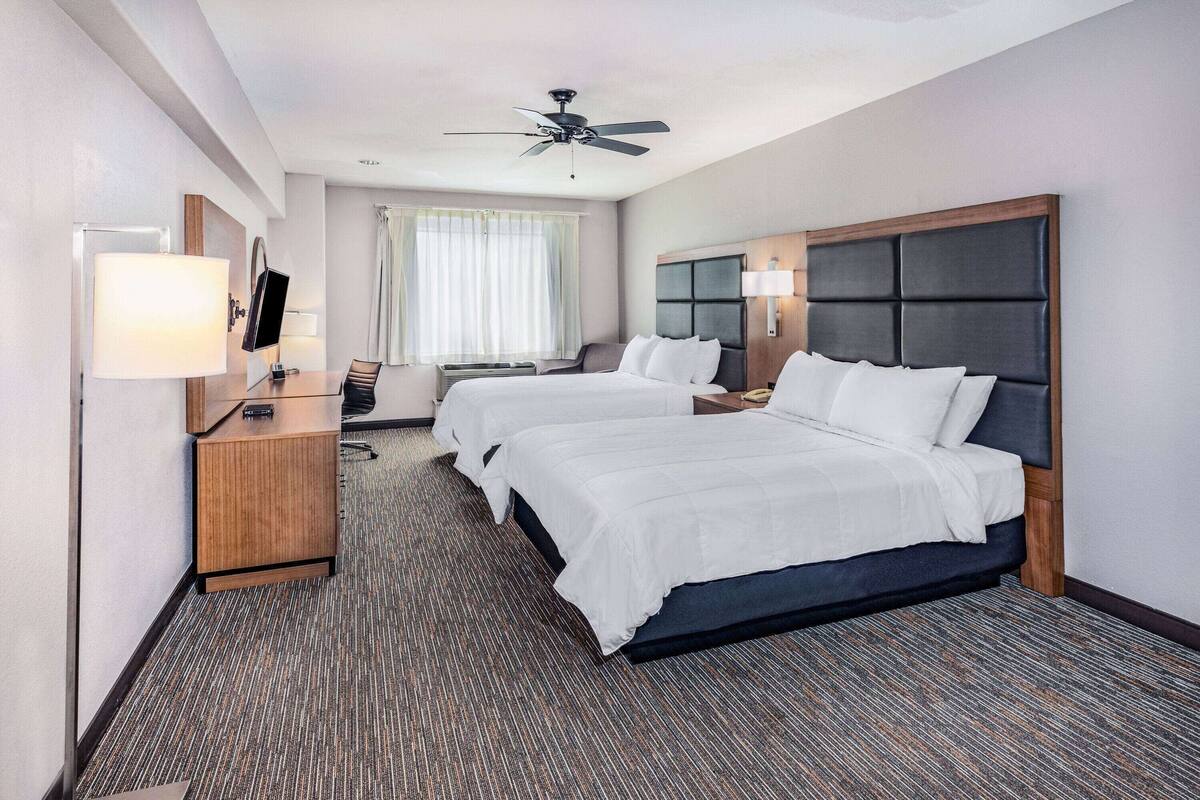 Une chambre d’hôtel avec deux lits, un bureau avec une lampe, un ventilateur de plafond et une fenêtre avec des rideaux.