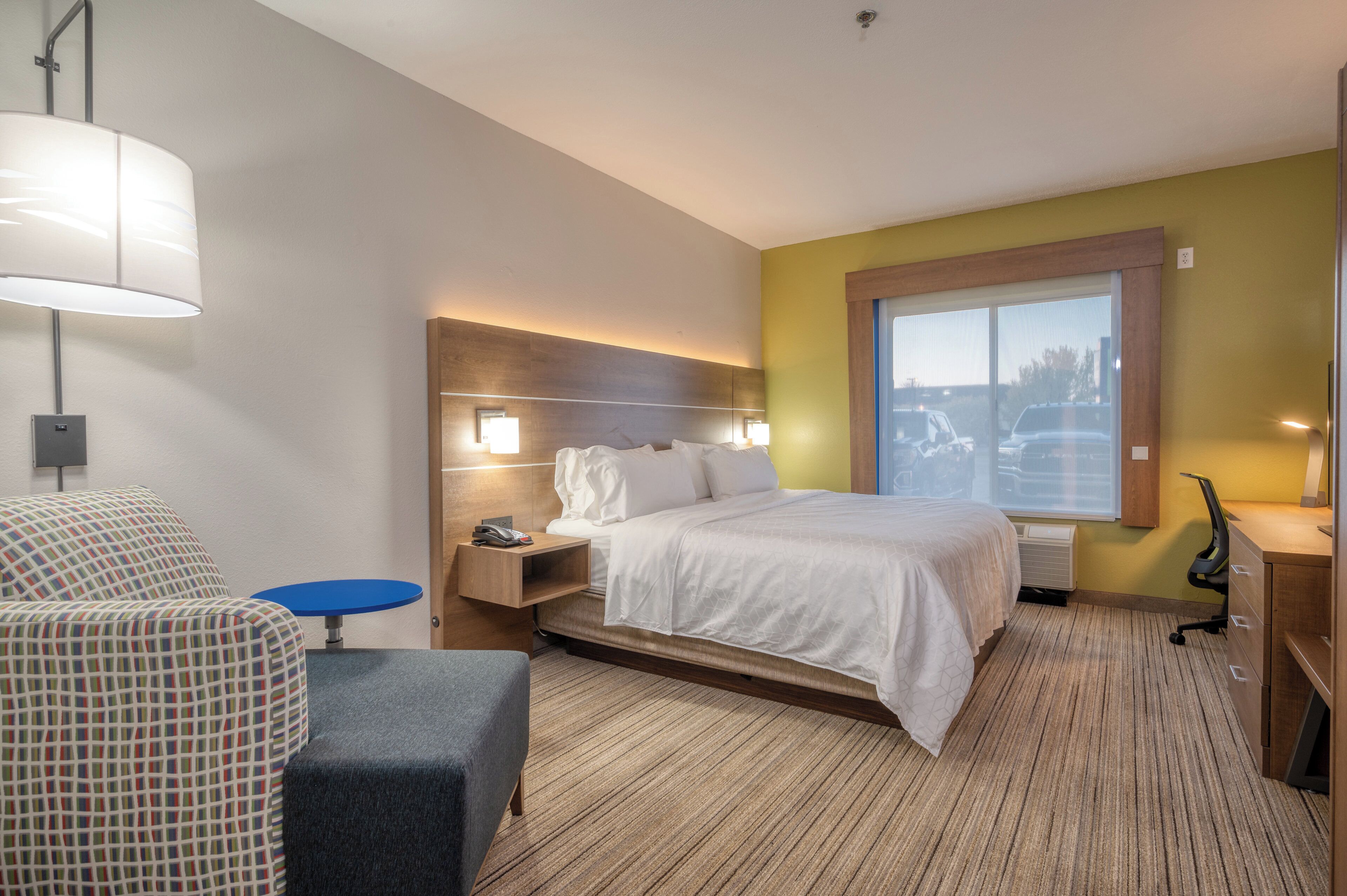 A hotel room with a bed, bedside table, lamp, chair, and a window with a view.
