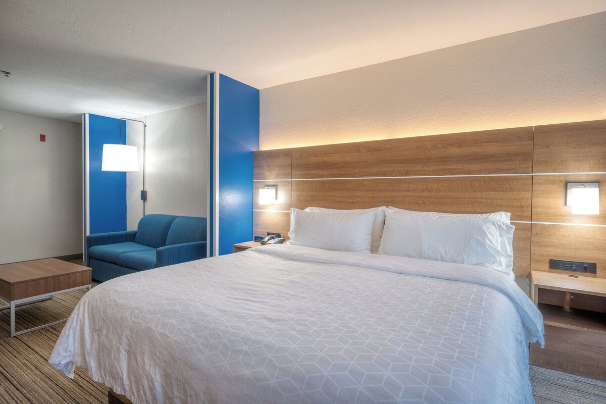 Ein Hotelzimmer mit einem großen Bett, einem blauen Sessel, einem Nachttisch mit Lampe und einem Holz-Kopfteil.