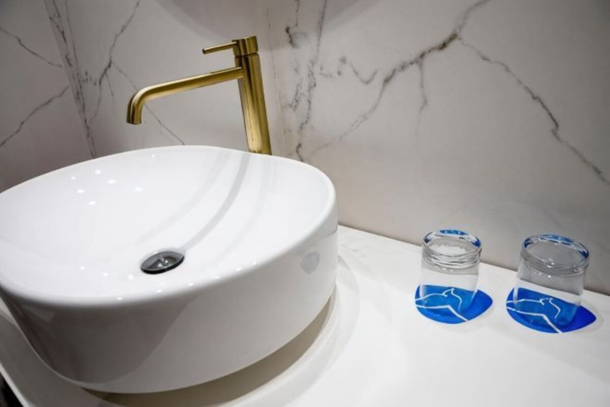 A white sink with a gold faucet, two small glass containers, and a marble wall.
