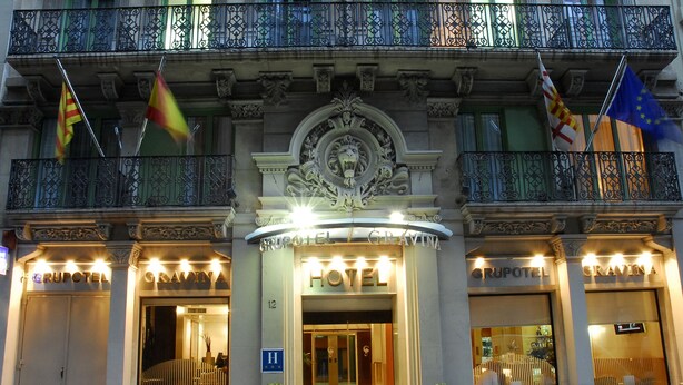A hotel facade with ornate architectural details, multiple flags, and illuminated signage. - Grupotel Gravina (Barcelona)