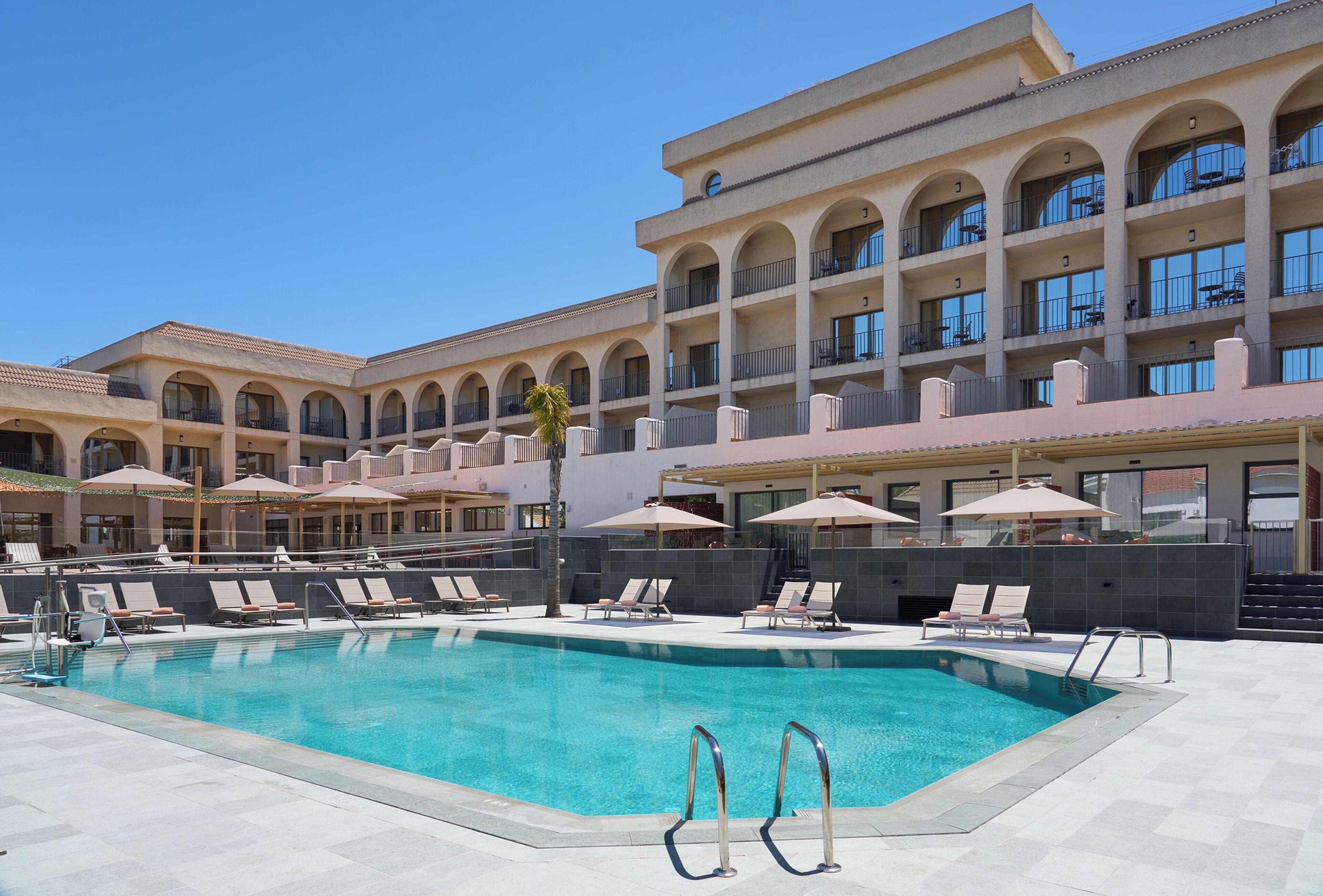 A pool area with lounge chairs and umbrellas, a clear blue pool, and a multi-story building with balconies.