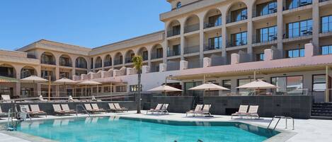 A pool area with lounge chairs and umbrellas, a clear blue pool, and a multi-story building with balconies.