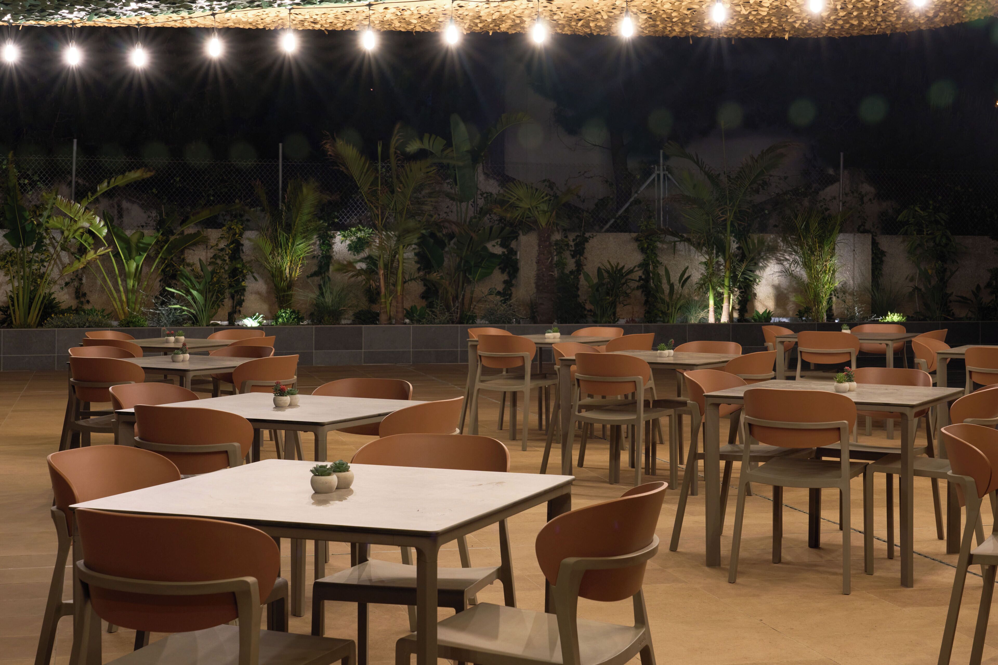 Un área de comedor al aire libre con varias mesas y sillas, rodeada de abundante vegetación y un techo decorativo.