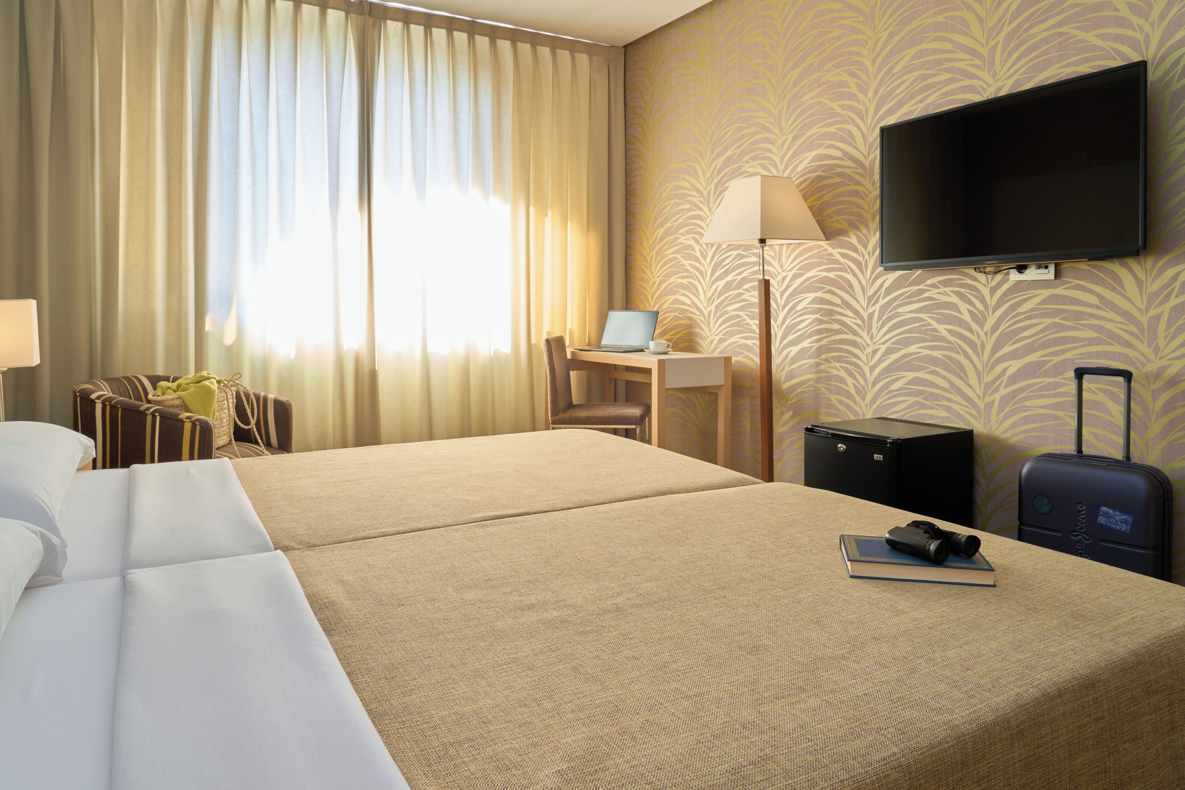 Habitación de hotel con una cama grande, un escritorio con una computadora portátil, un televisor y una maleta.