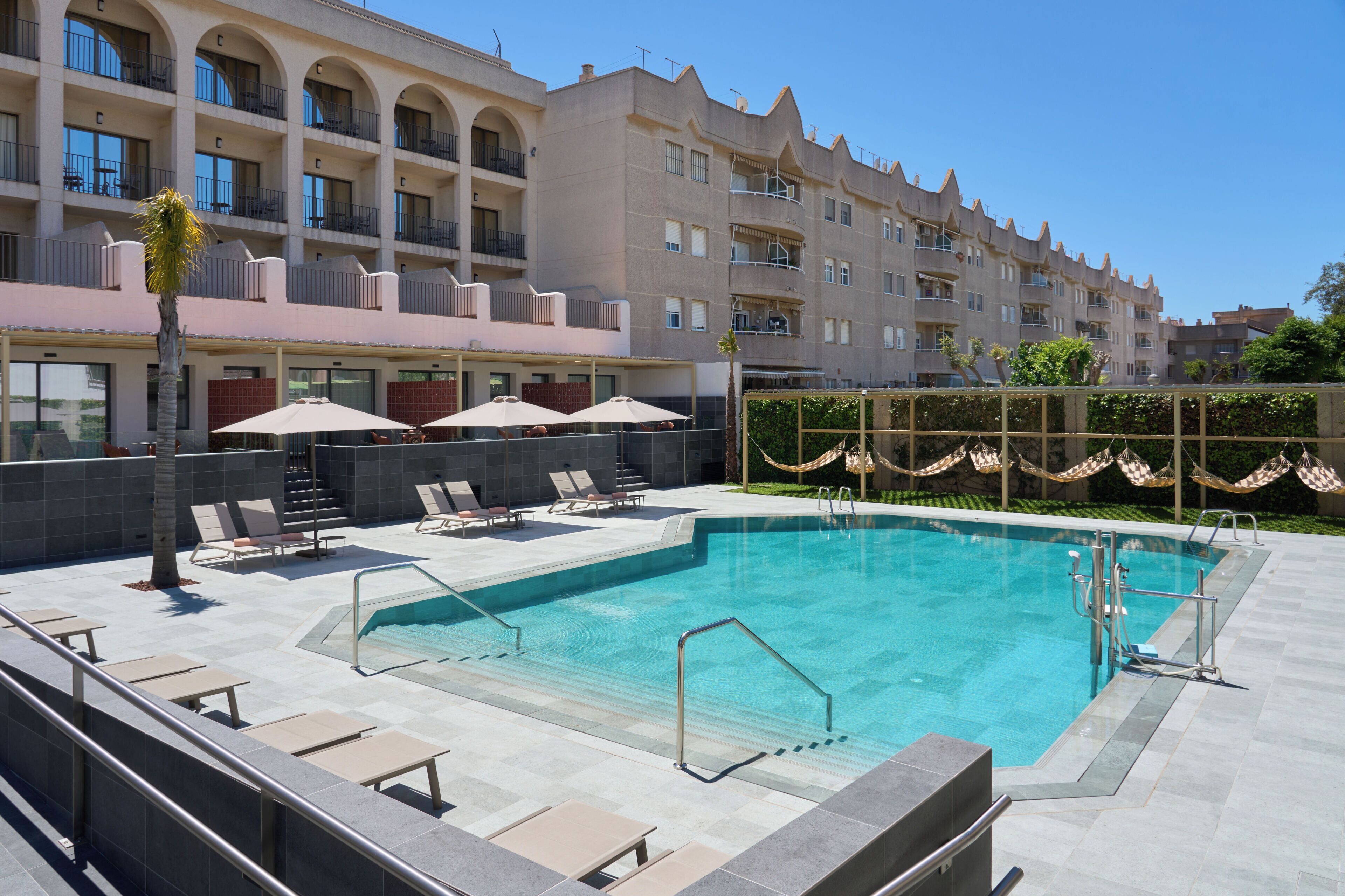 Une piscine entourée de chaises longues et de parasols, elle-même bordée d’un immeuble à plusieurs étages doté de balcons.