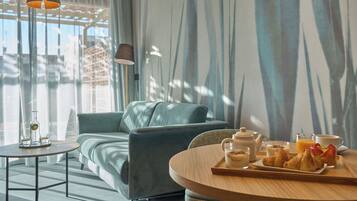 A modern living room with a sofa, a round table with breakfast items, and a large window with a view of buildings.