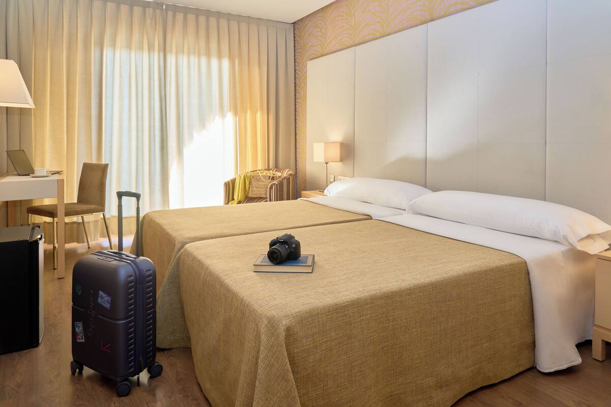 Una habitación de hotel con dos camas, un escritorio, una silla, una lámpara y una maleta.