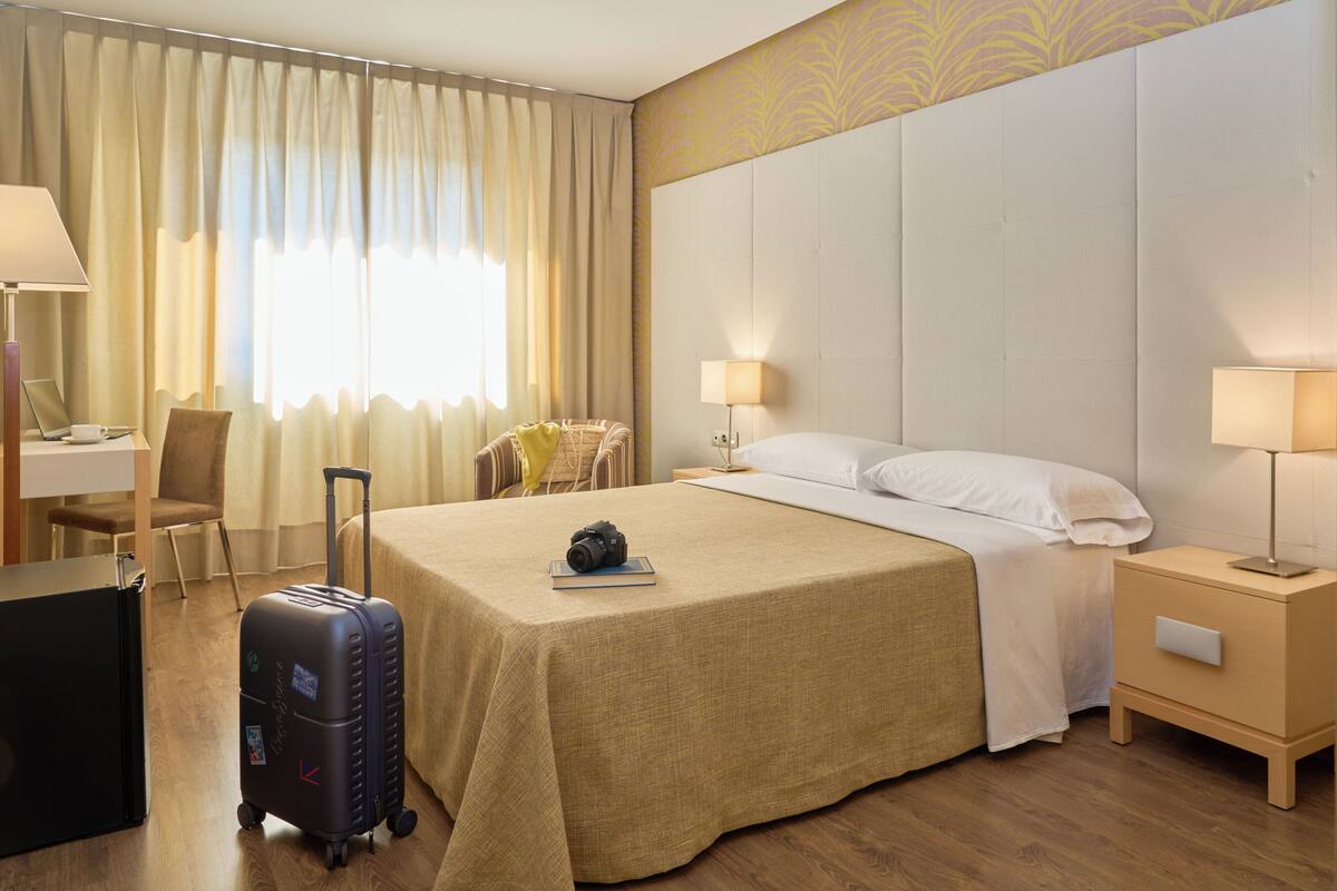 Habitación de hotel con cama, escritorio, silla, maleta y mesita de noche con lámpara.