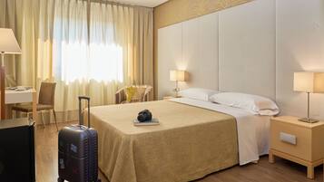 A hotel room with a bed, a desk, a chair, a suitcase, and a nightstand with a lamp.