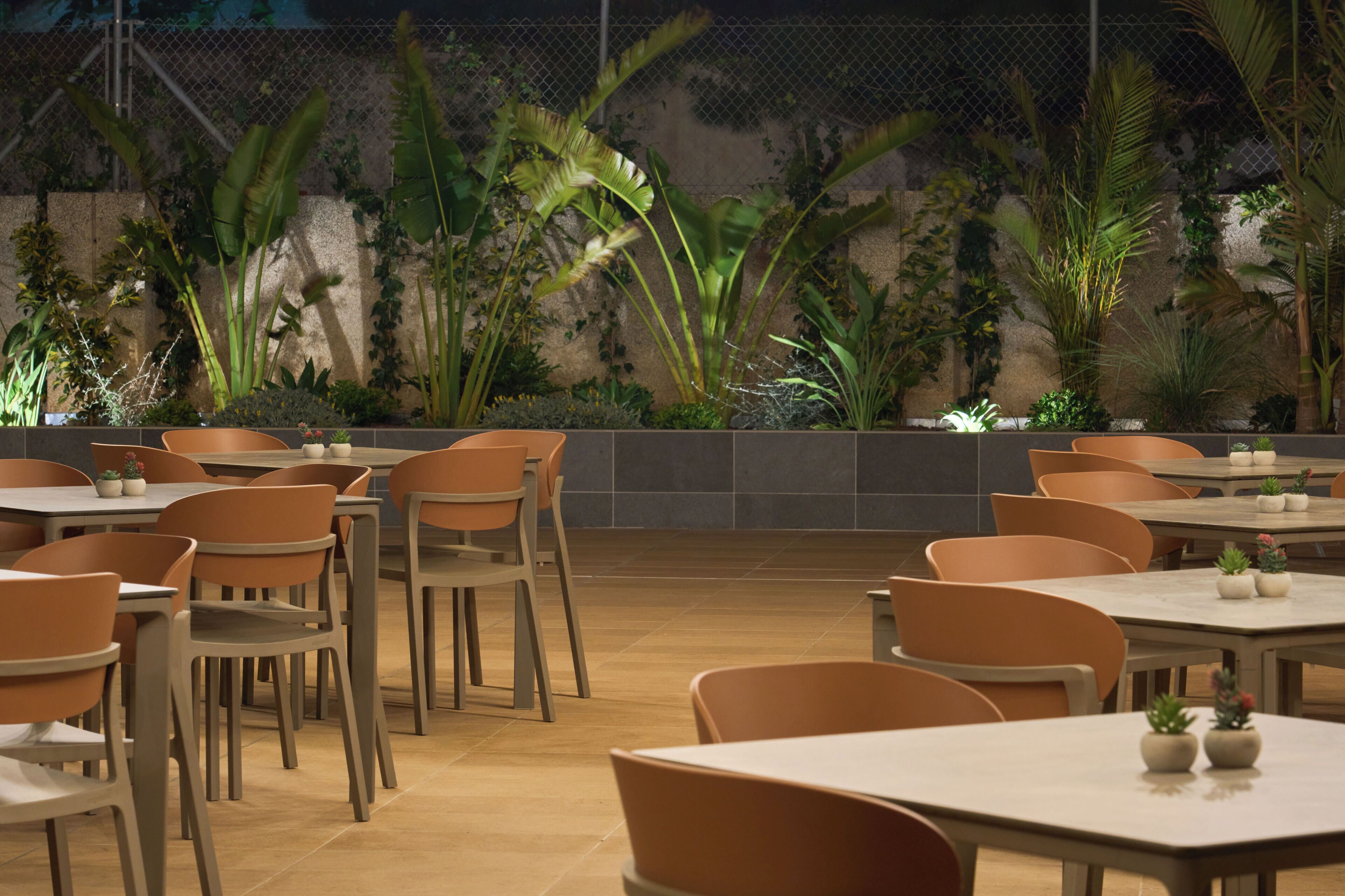 An indoor dining area with multiple tables and chairs, surrounded by a variety of plants and a stone wall.