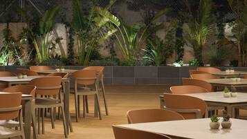 An indoor dining area with multiple tables and chairs, surrounded by a variety of plants and a stone wall.