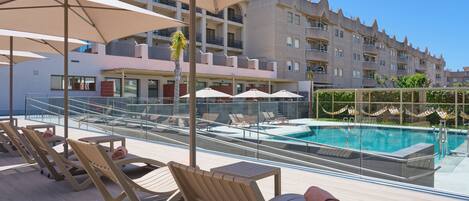 A rooftop pool area with lounge chairs and umbrellas, a clear view of the pool, and a modern building in the background.