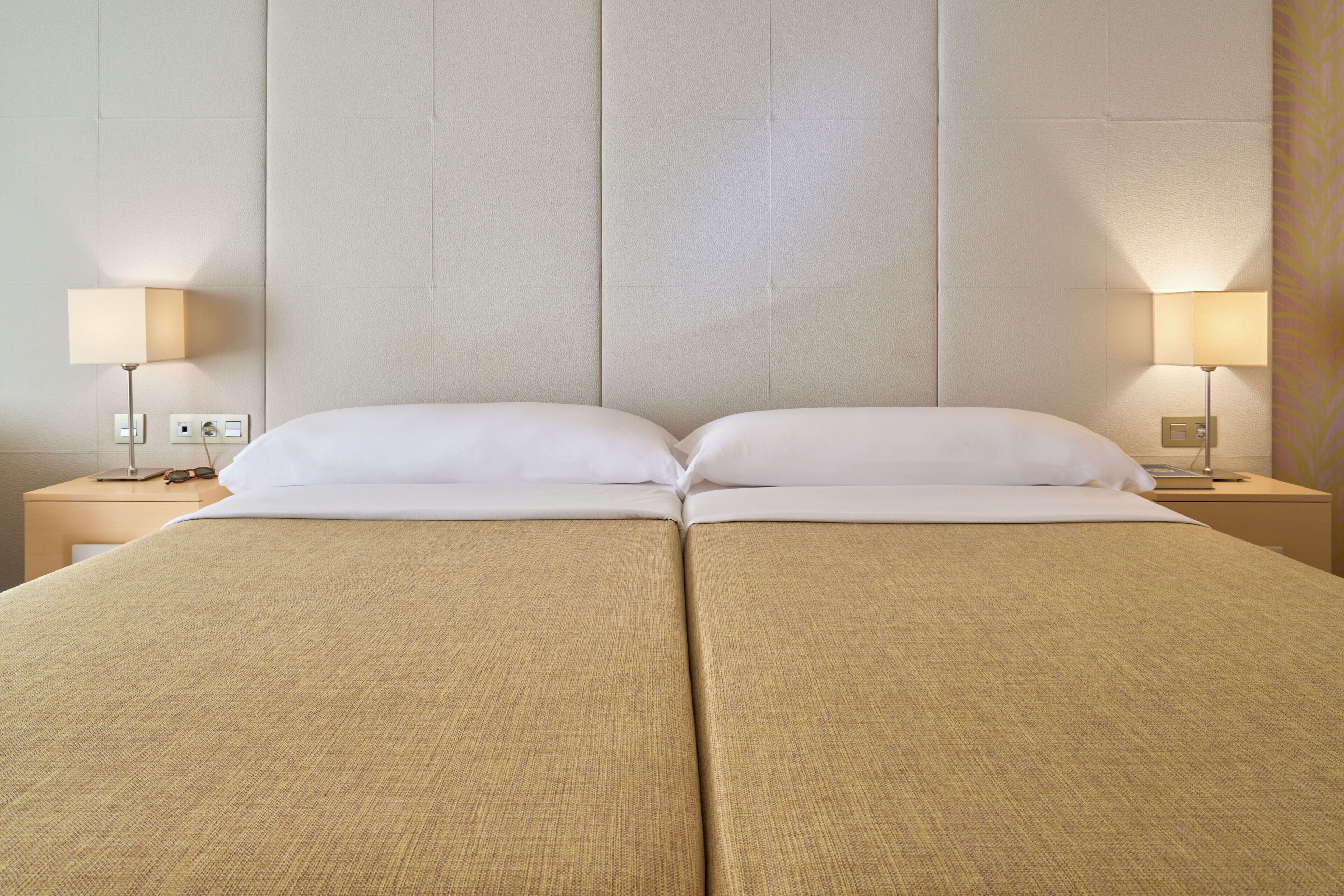 Ein Doppelbett mit beiger Bettdecke, zwei Nachttische mit Lampen und eine schlichte Wand mit dezentem Muster.