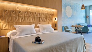 Una habitación de hotel moderna con una cama grande, cabecera de madera, una mesita con lámpara y una zona de estar con sofá y sillones.