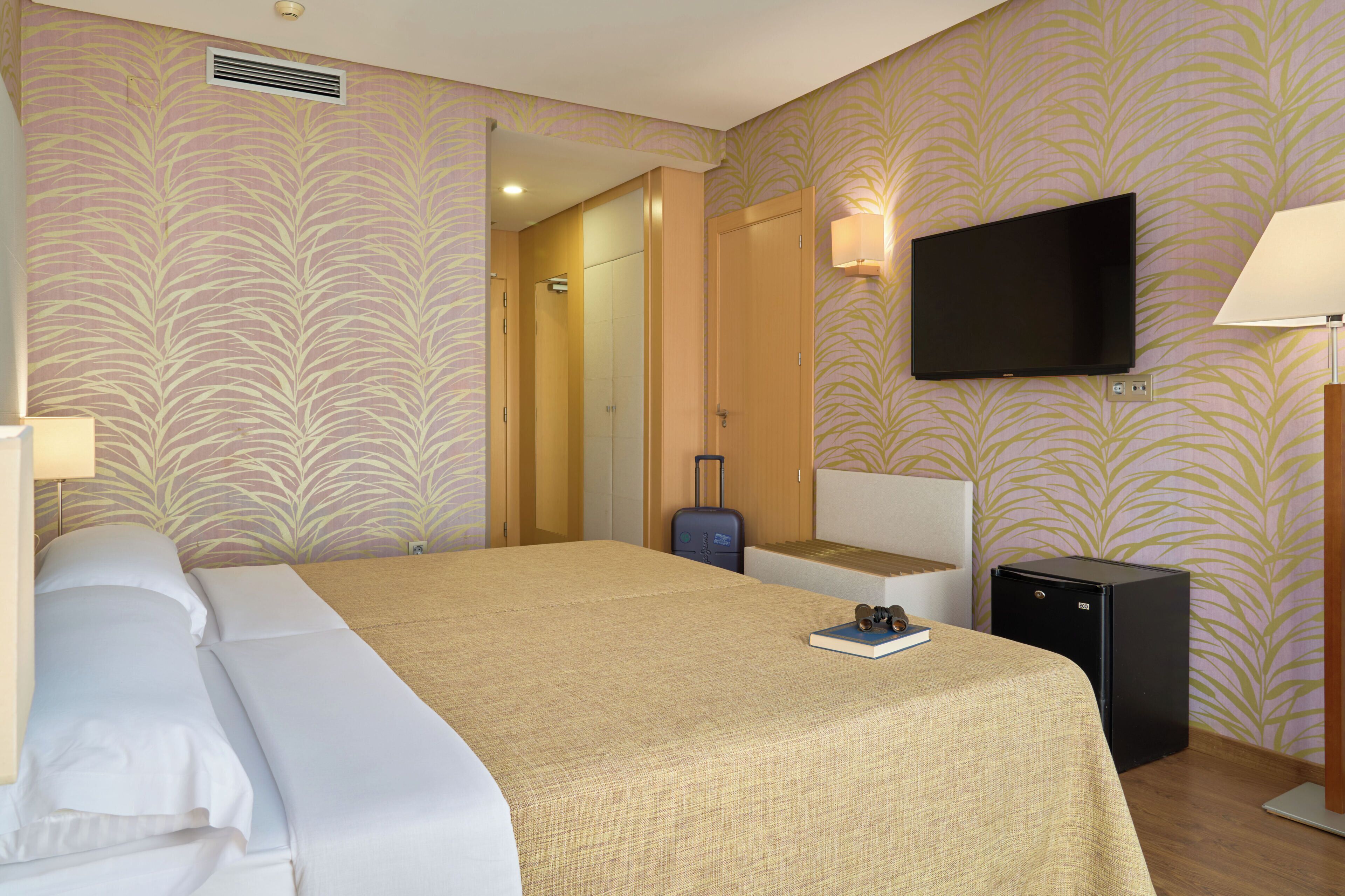 A hotel room with a bed, a television, a desk, a suitcase, and a compact refrigerator.