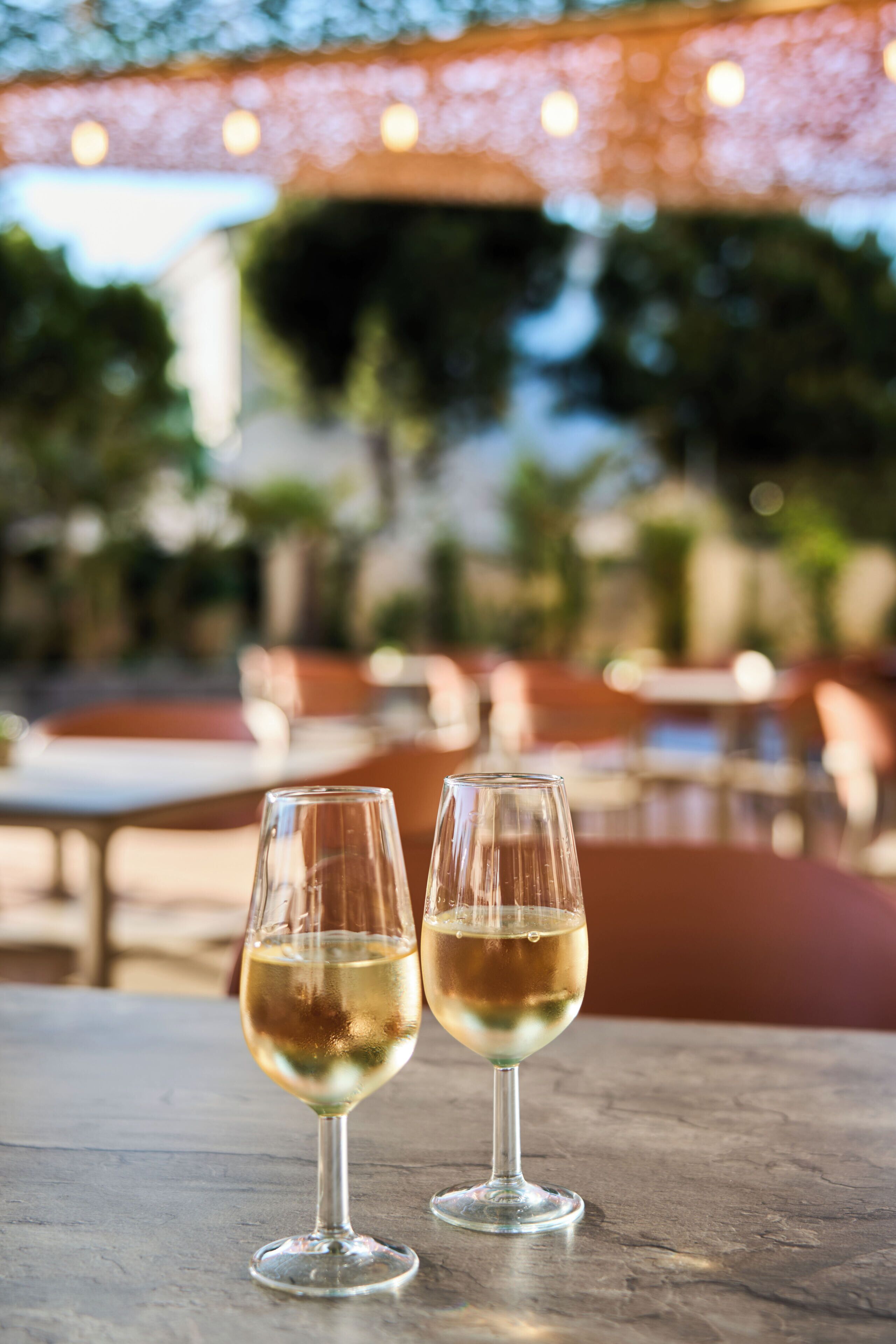 Two glasses of white wine on a table with a blurred outdoor seating area in the background.