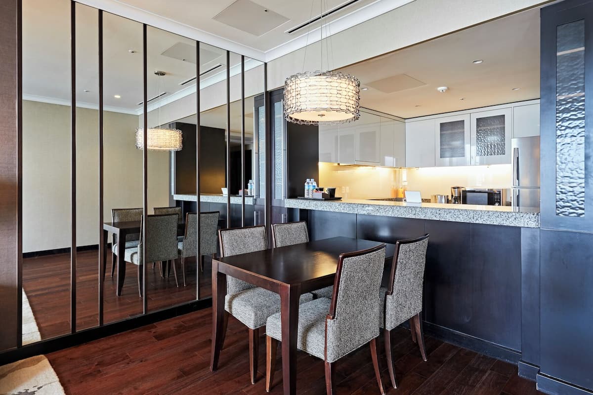 A modern dining area with a dark wooden table and chairs, a glass partition, and a kitchen with stainless steel appliances.
