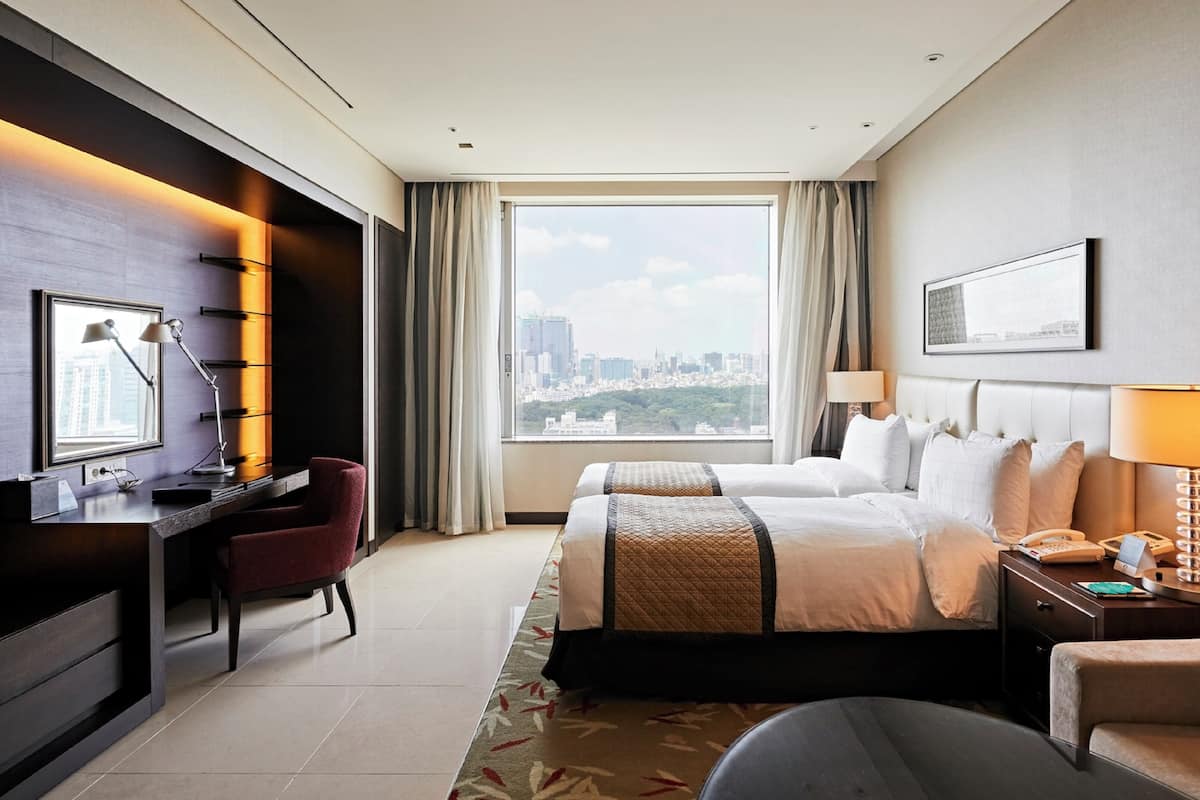 A hotel room with a desk, two beds, a chair, a nightstand, and a view of the cityscape.