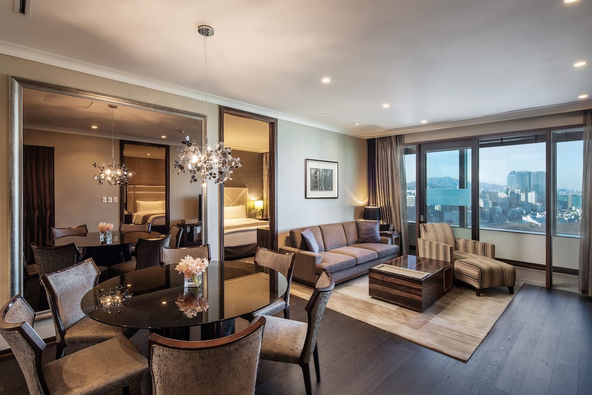 A modern hotel room with a dining area, a sofa set, and a large window offering a city view.