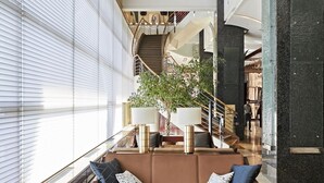 Un vestíbulo amplio con una zona de estar central que incluye un sofá marrón y una mesa de centro de madera. Una escalera con barandilla de cristal conduce a un nivel superior.