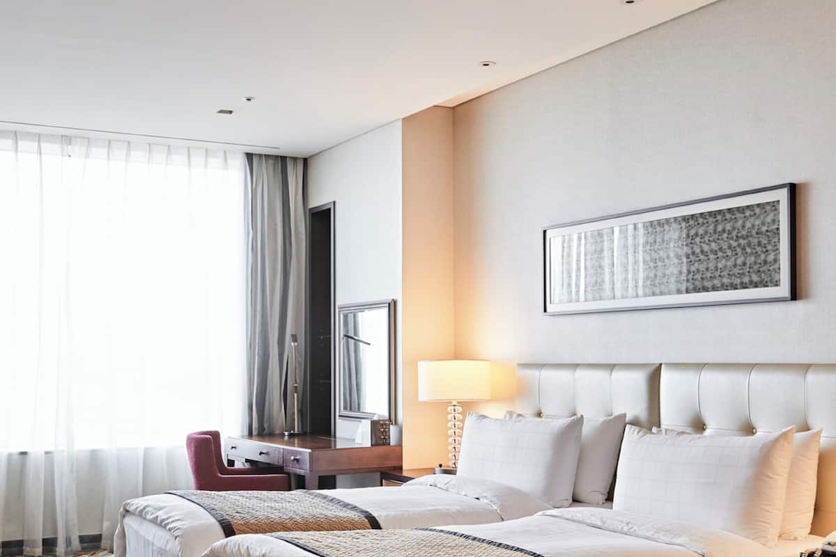 A hotel room with two beds, a desk, a chair, a lamp, and a large window with curtains.