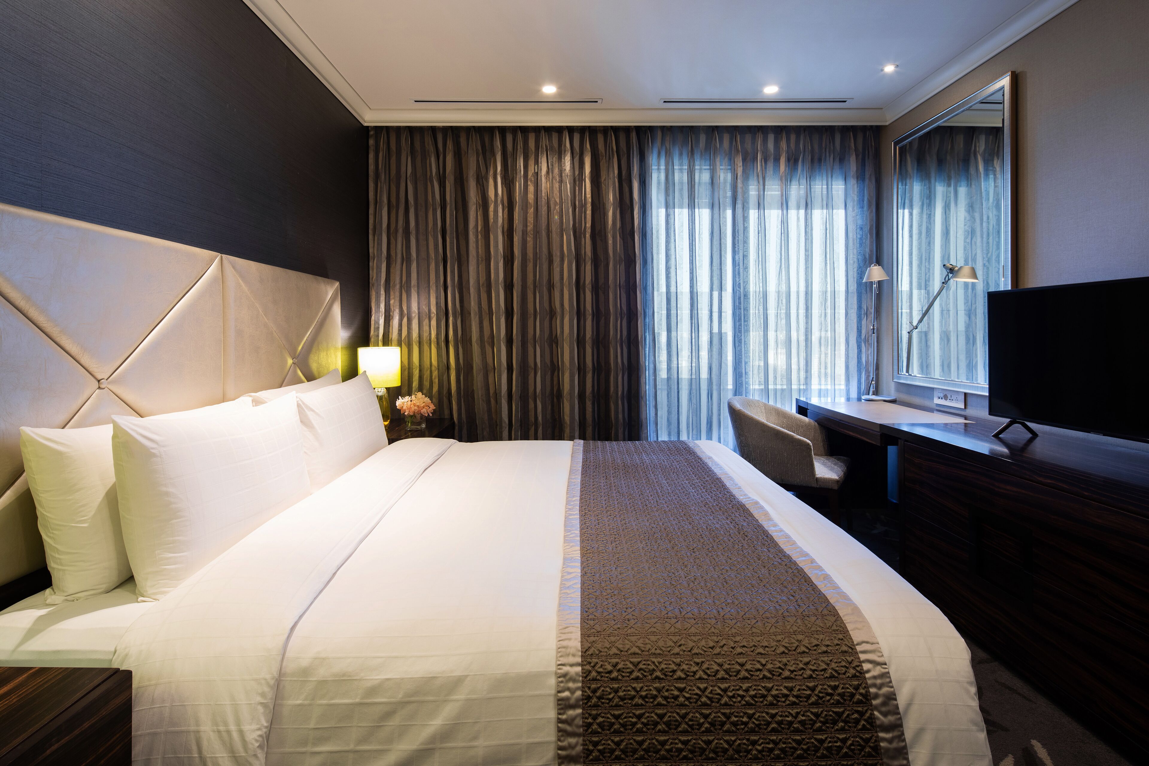 Une chambre d’hôtel avec un grand lit, un bureau, une chaise, une télévision et une grande fenêtre avec des rideaux.