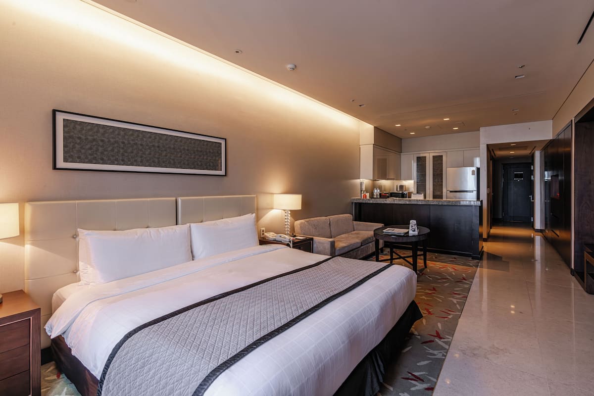 A modern hotel room with a large bed, a sitting area, a kitchenette, and a bathroom.
