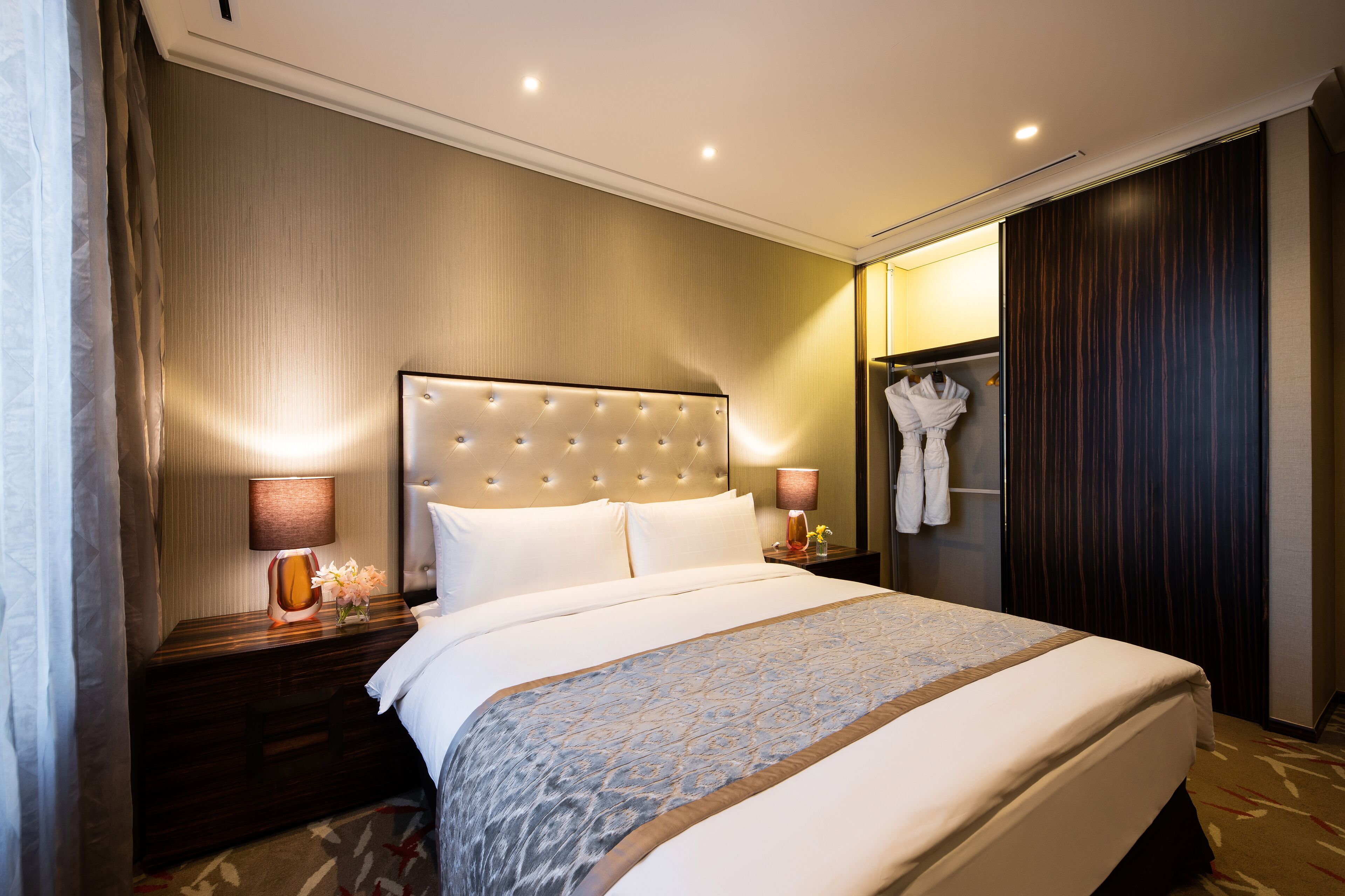 A hotel room with a large bed, a nightstand with a lamp, a robe hanging on the wall, and a dark wooden wardrobe.