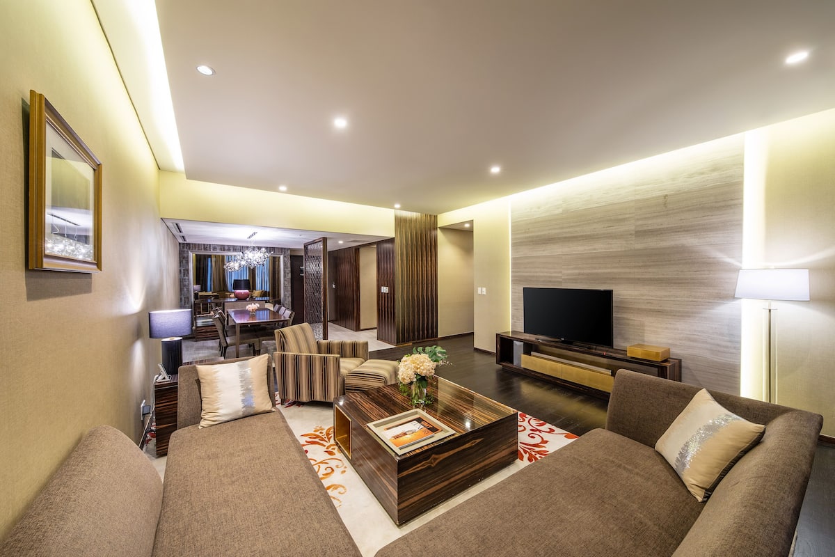 A modern living room with a sofa, coffee table, and TV. There is a dining area in the background.
