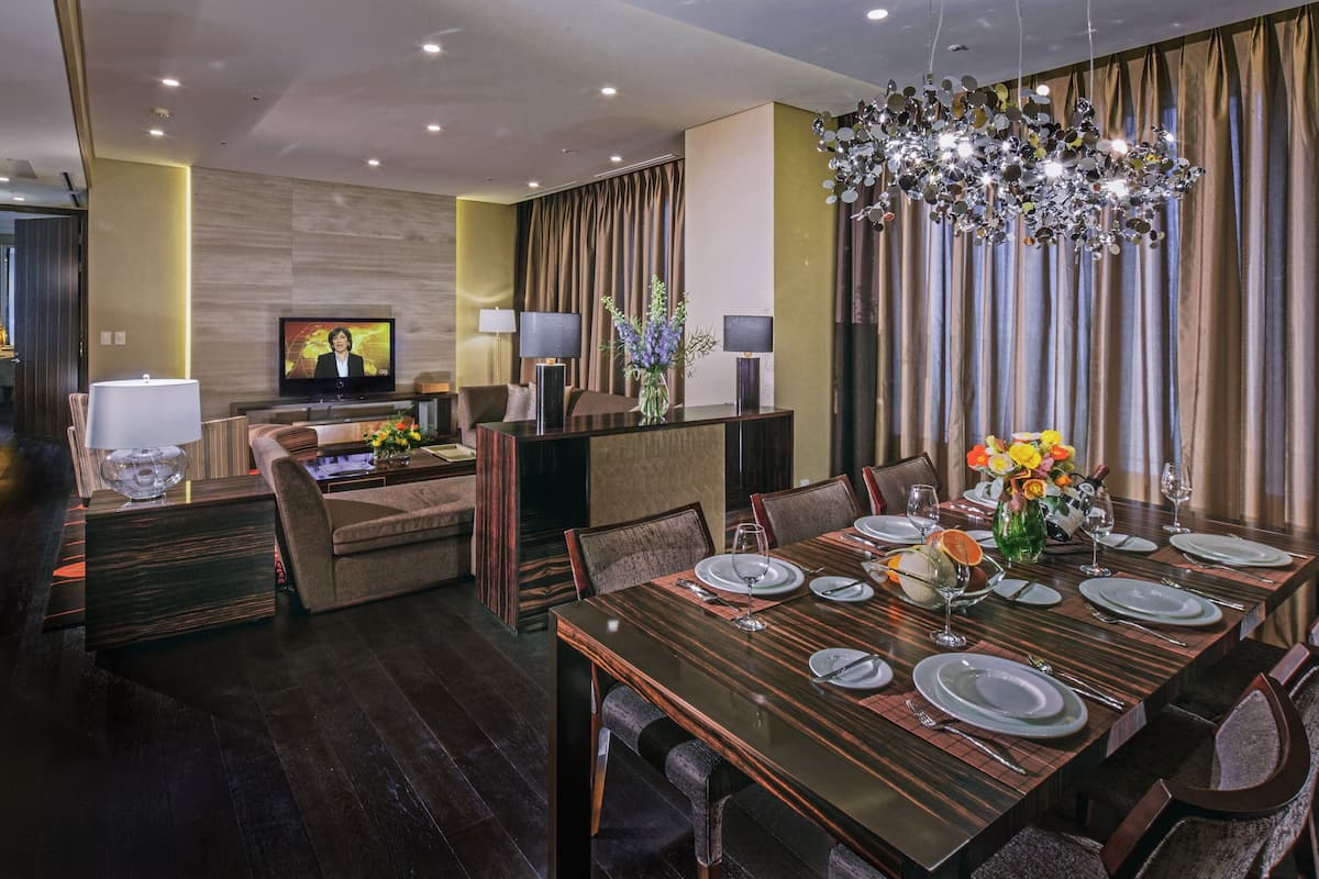 A modern dining area with a wooden table set for six, a television in the background, and a decorative chandelier.
