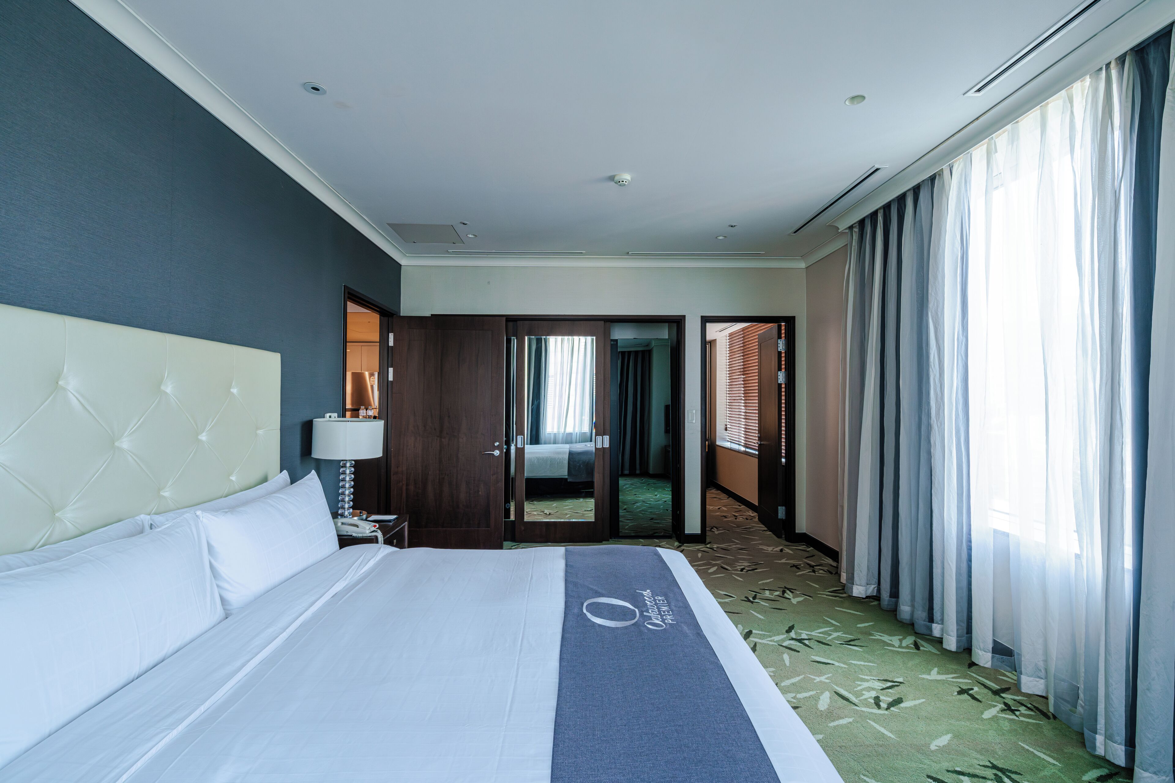 A hotel room with a bed, a nightstand, a lamp, a door leading to another room, and a view of a hallway.