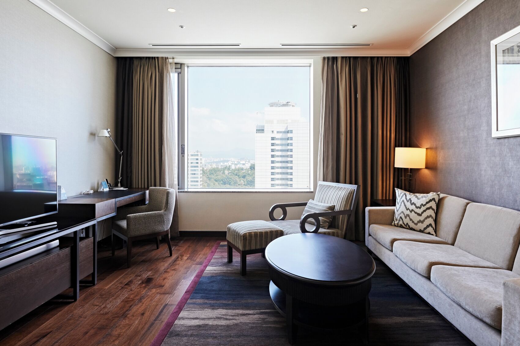 A hotel room with a large window, a flat-screen TV, a desk, a sofa, and a coffee table.
