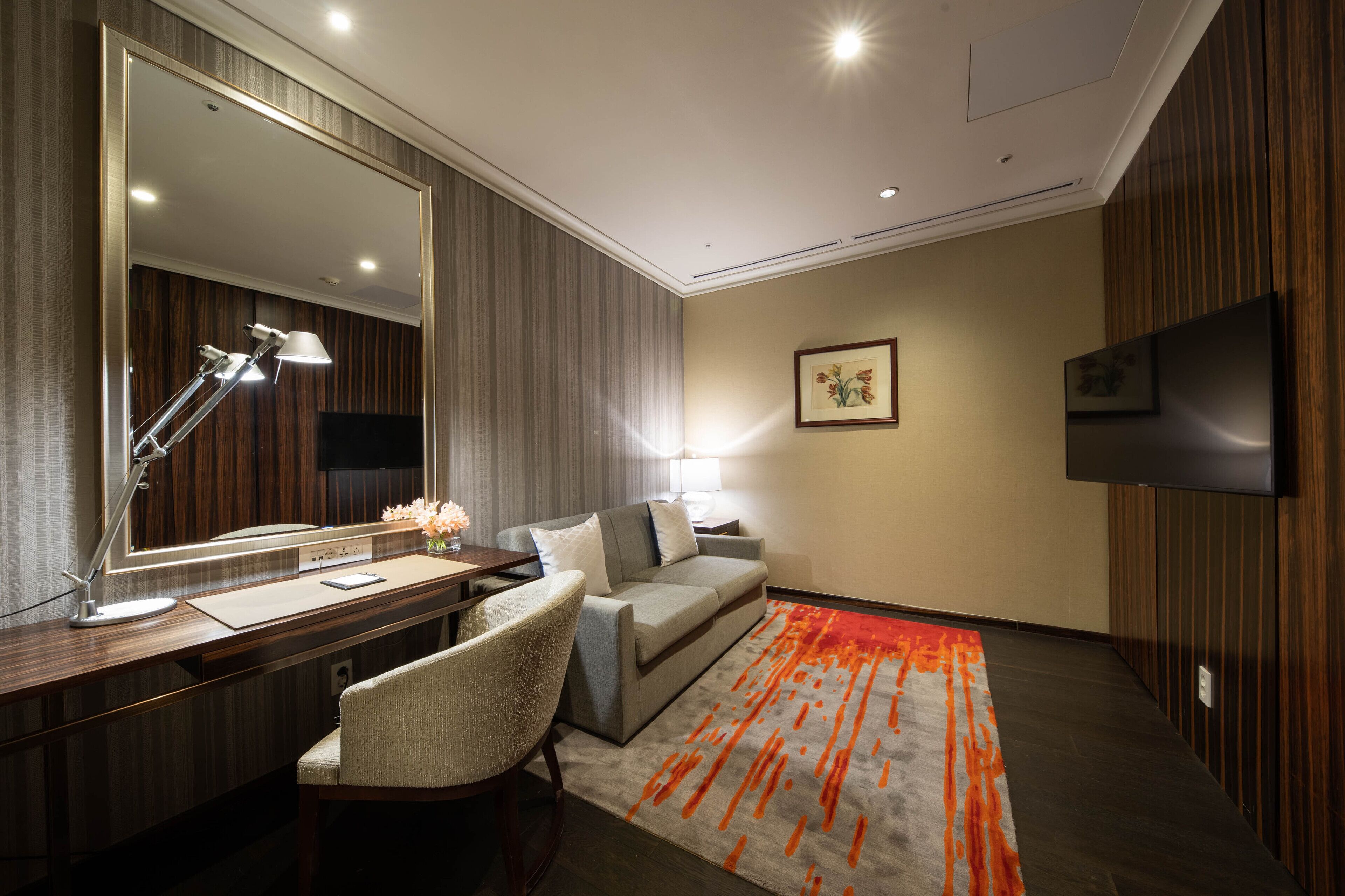 A hotel room with a large mirror, a desk with a lamp, a sofa, a TV, and a patterned rug.