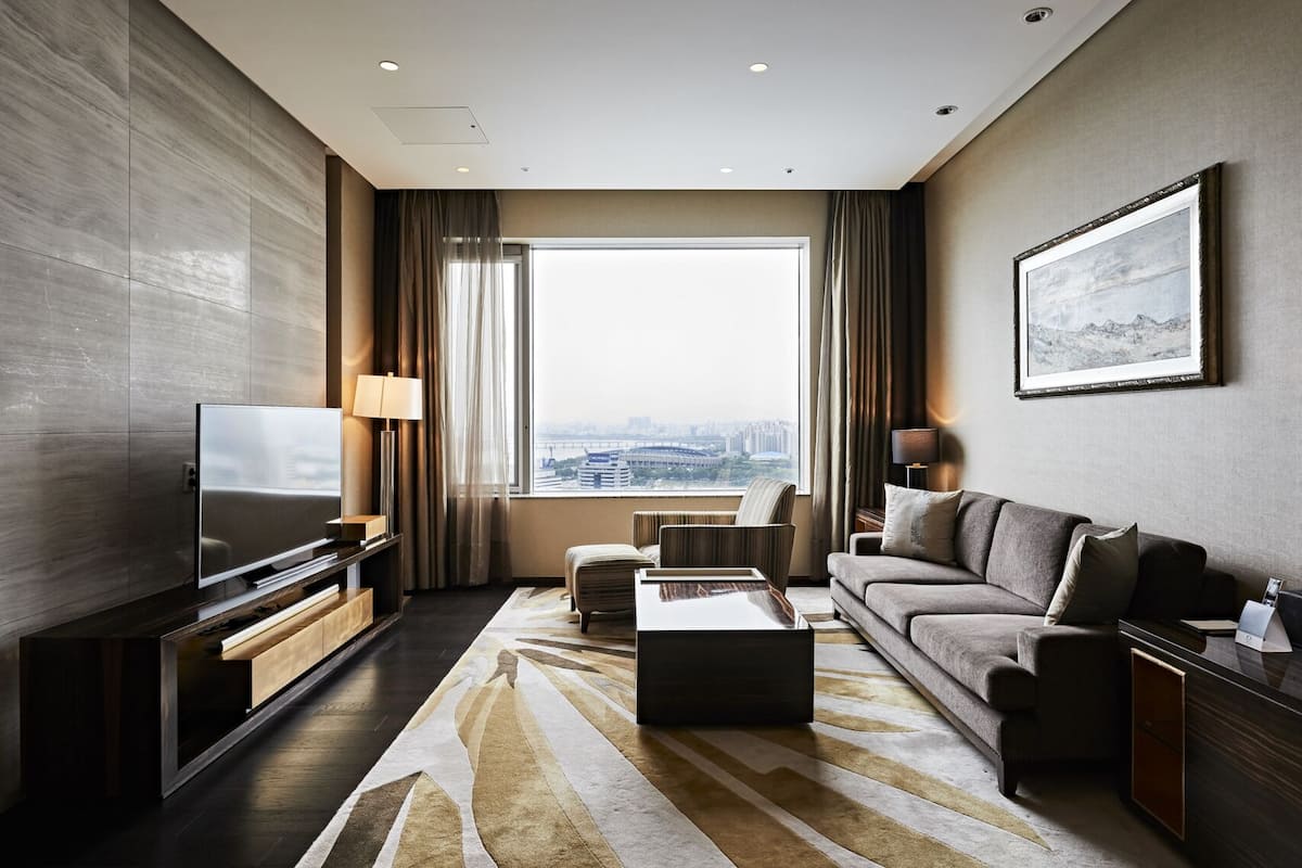 A modern hotel room with a large window, a flat-screen TV, a sofa, a coffee table, and a framed picture on the wall.