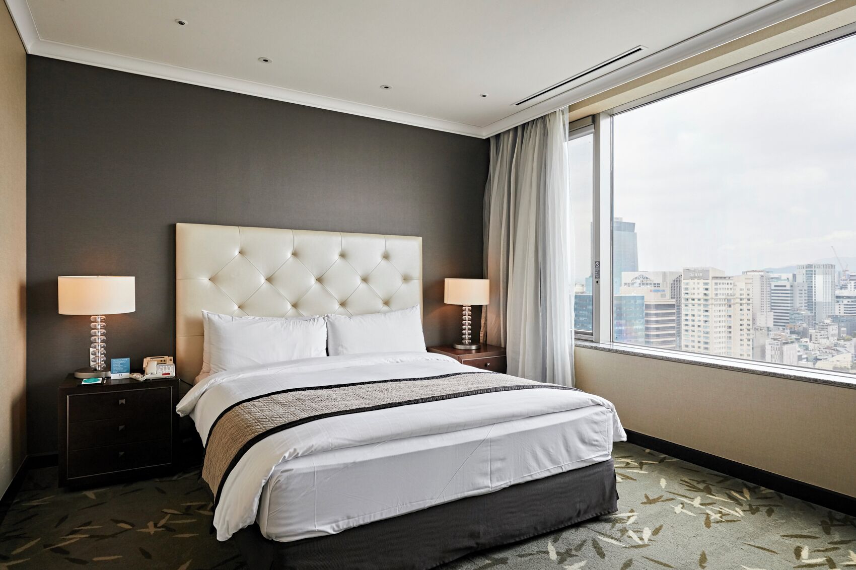 Habitación de hotel con una cama grande, dos mesitas de noche con lámparas, vistas a la ciudad y una pared de color oscuro.